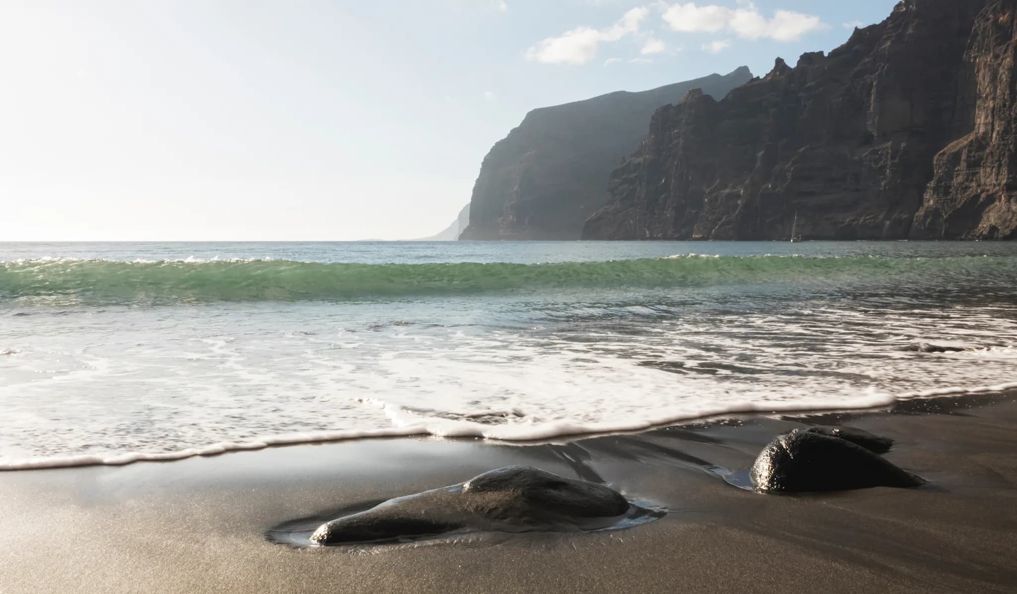 Waves washing over dark volcanic sand and smooth black stones, with sea cliffs in the background