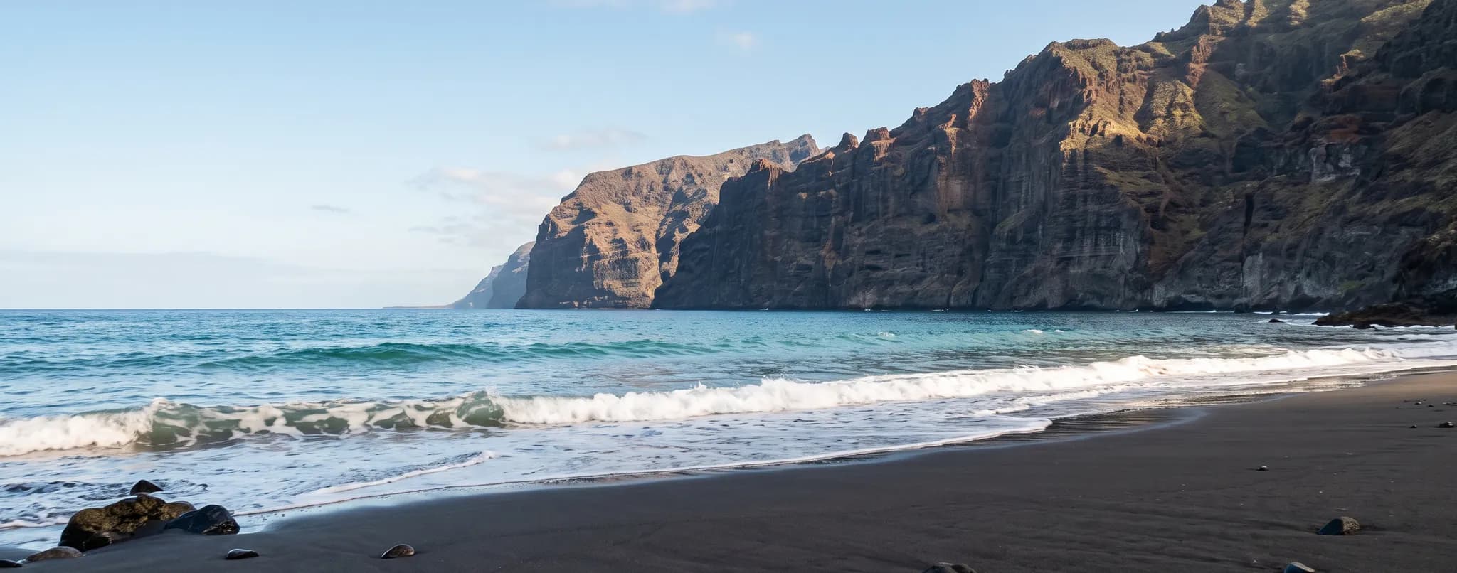 Black sand beach on Tenerife beneath towering volcanic cliffs, with Atlantic waves breaking on the shore
