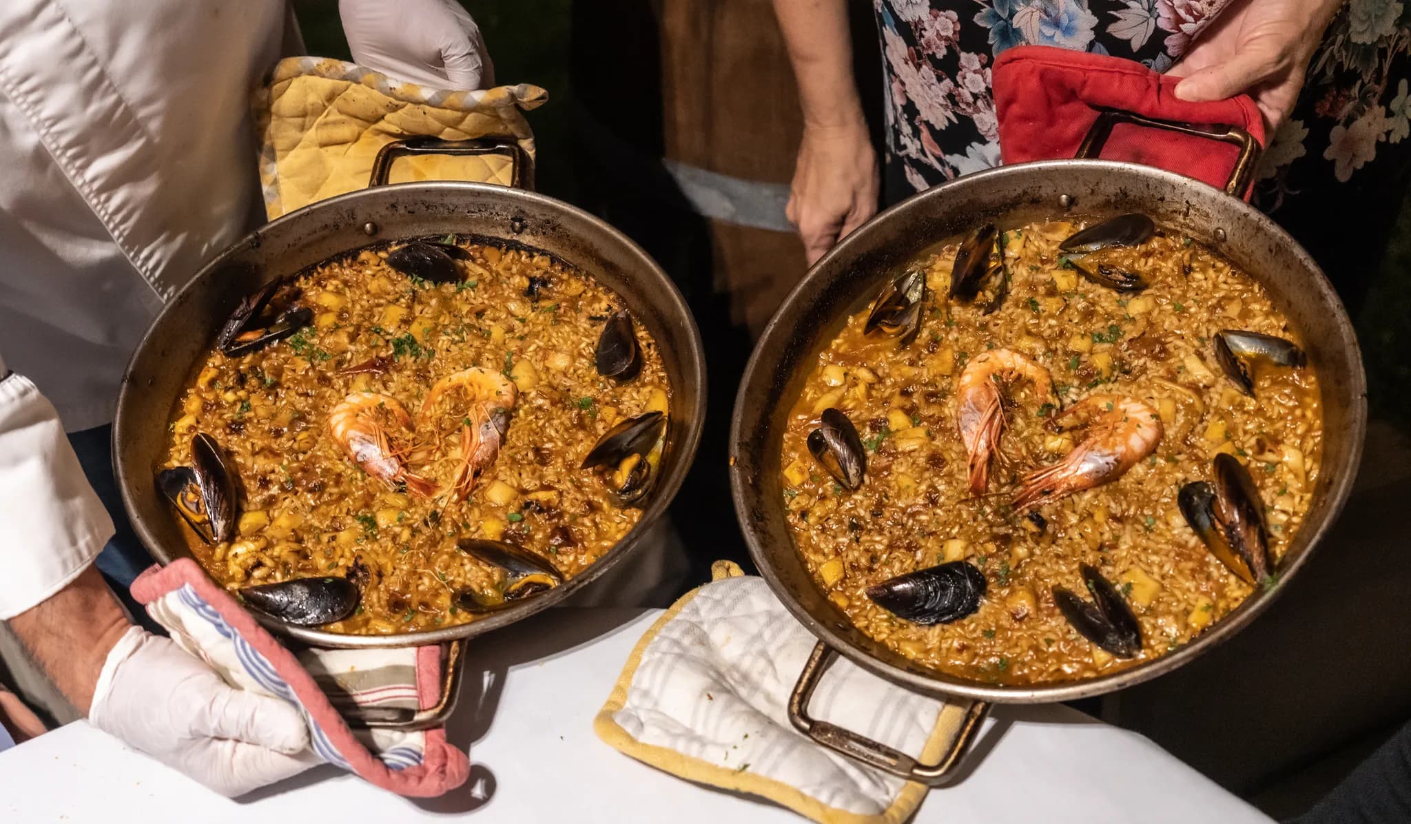 Two cooks holding up freshly made seafood paellas in their pans