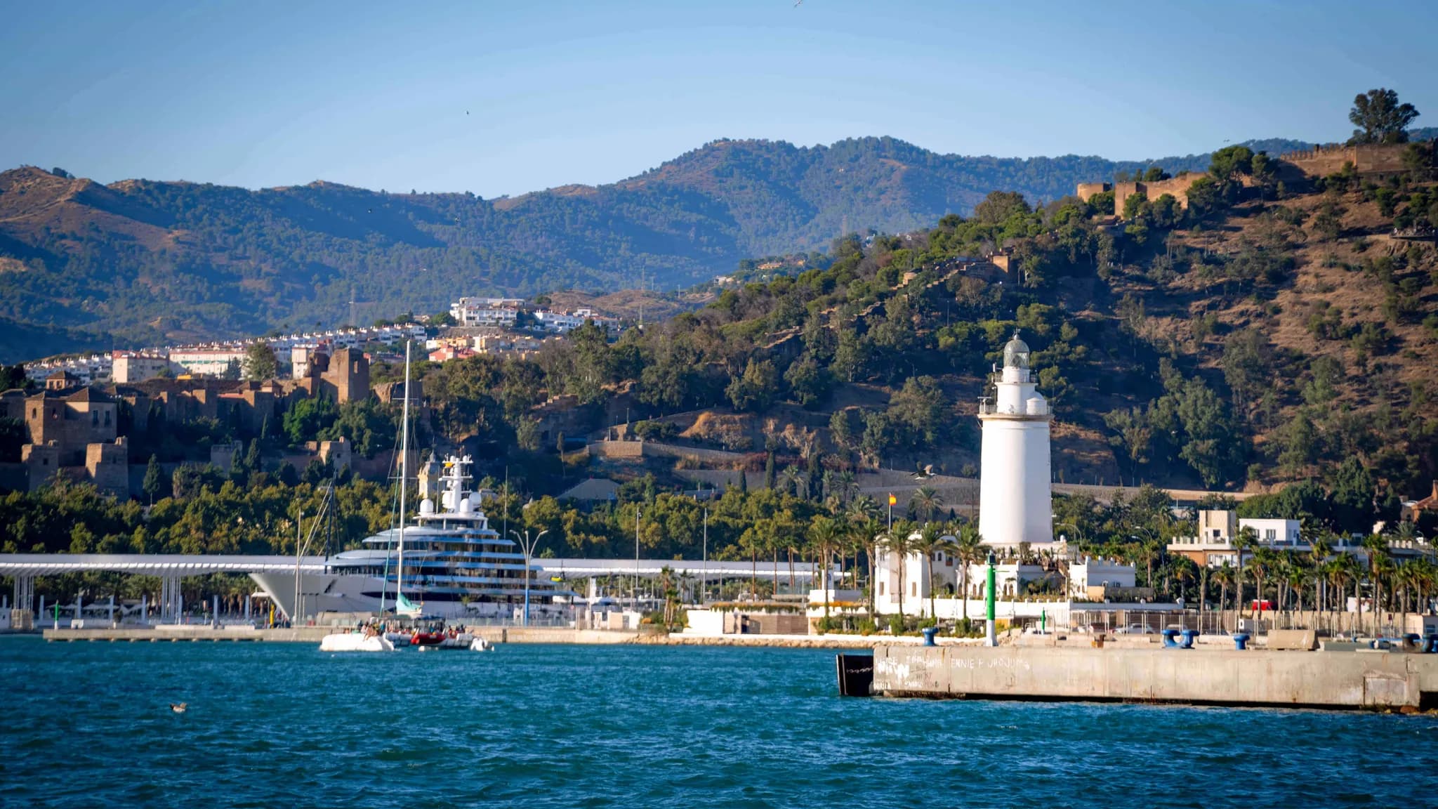 Malaga harbour with La Farola lighthouse, a superyacht at the quay and the Alcazaba on the wooded hillside behind