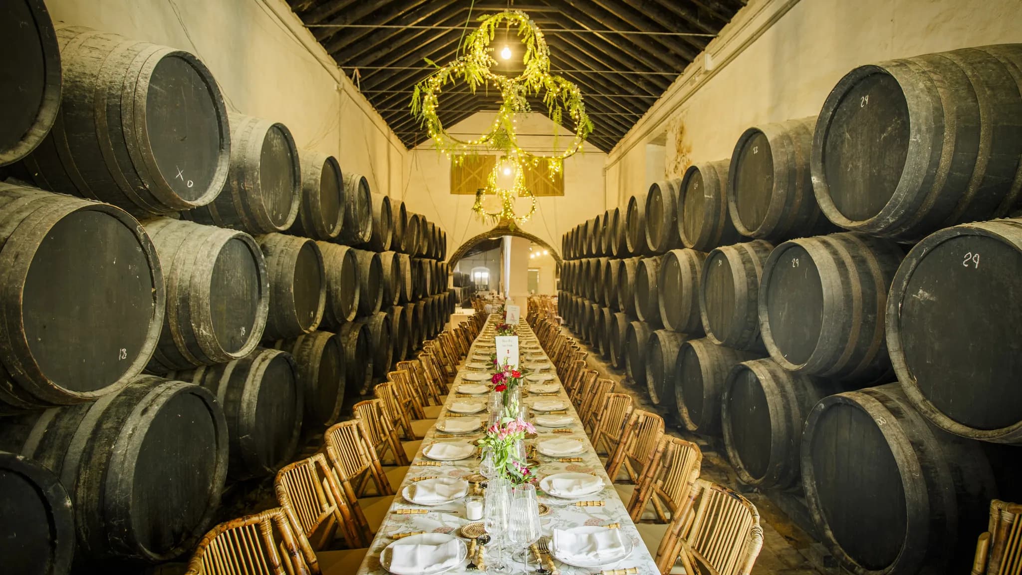 A long banquet table set between rows of barrels in a wine cellar, beneath a floral chandelier