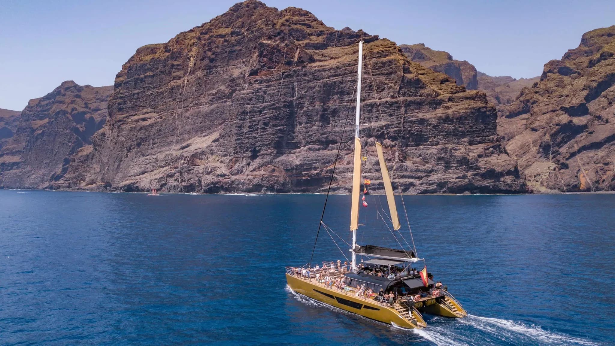 A sailing catamaran full of guests cruising in front of towering sea cliffs